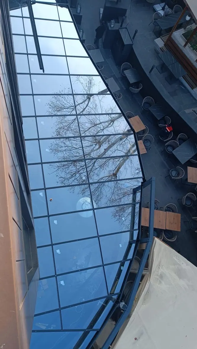Glasdach einer Terrassenüberdachung von oben, darin spiegeln sich Bäume und Himmel